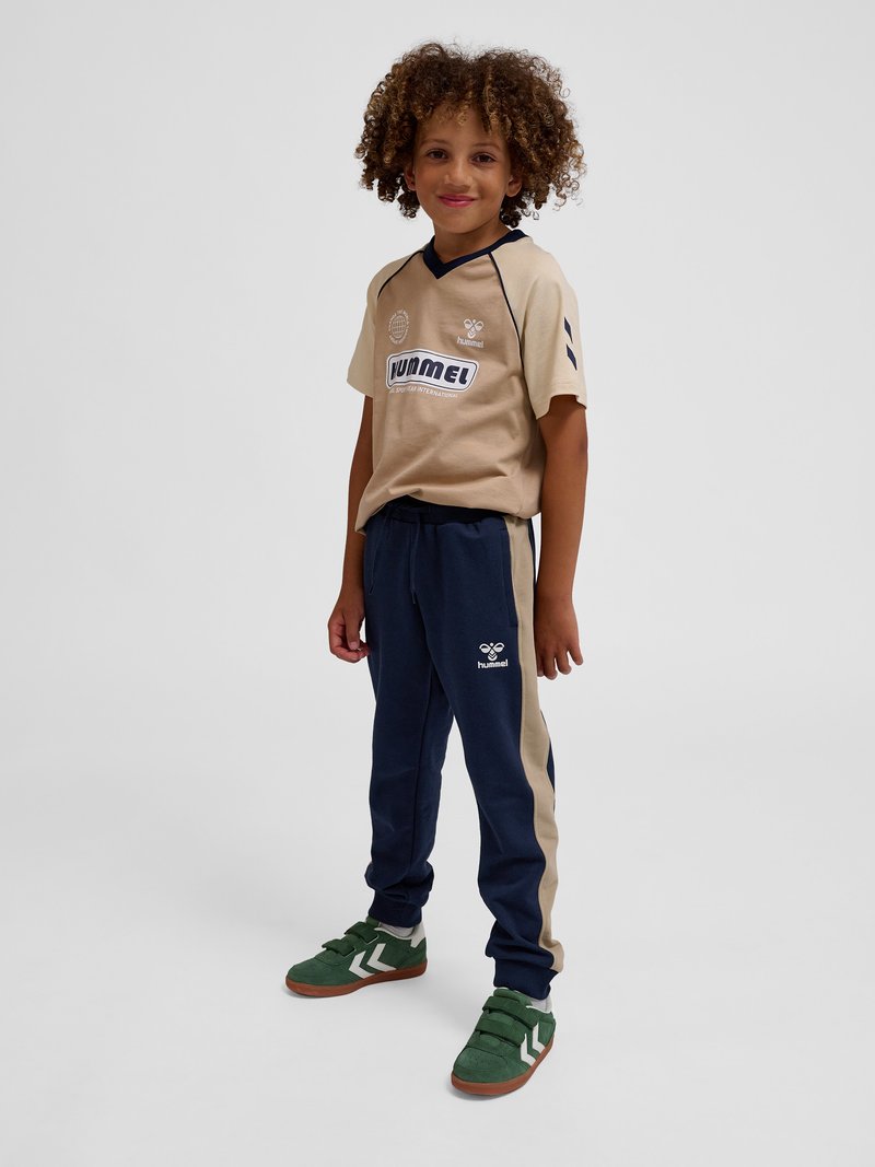 Dziecko o kręconych włosach, ubrane w beżową i granatową odzież sportową oraz zielone trampki, stojące na jednolitym jasnym tle.