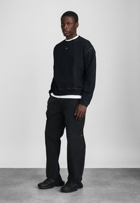 Crna dukserica s okruglim izrezom i vidljivim detaljima šivanja, uparen s tamnim trapericama i robusnim crnim tenisicama. Jednostavan, ležeran izgled.