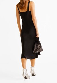 Robe noire sans manches avec une silhouette ajustée, longueur mi-mollet et texture lisse ; associée à des bottines argentées et un sac à motifs.