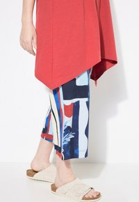 Tunic asimmetrica rossa, pantaloni cropped con motivo in blu, rosso e crema, abbinati a sandali beige con due cinturini regolabili.