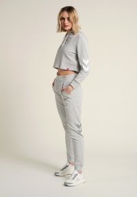 Grå kort sweatshirt med vitt chevronmönster på ärmarna, kombinerad med grå joggers med sidofickor och matchande chevrondetalj. Vita sneakers.