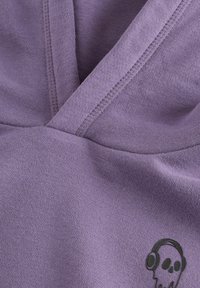 Gros plan sur un tissu violet avec des coutures détaillées et un petit crâne noir portant des écouteurs imprimé près du coin inférieur droit.