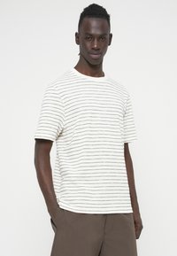 Λευκό κοντομάνικο t-shirt με μαύρες οριζόντιες ρίγες, στρογγυλή λαιμόκοψη και απαλή υφή. Συνδυάζεται με σκούρο καφέ παντελόνι.