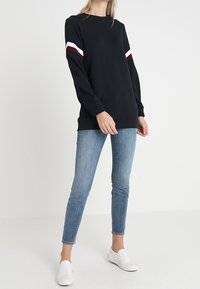 Persona que lleva una sudadera negra de manga larga con rayas blancas y marrones en las mangas, jeans ajustados azules y zapatillas blancas sin cordones.