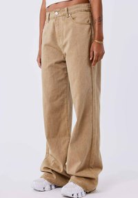 Vaqueros de pierna ancha beige de denim, con cintura alta, cierre de botón y costuras laterales sutiles. Llevados con zapatillas blancas.
