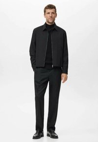 Chaqueta negra con cremallera y corte recto, llevada sobre un suéter negro de cuello alto, combinada con pantalones negro de pierna recta y zapatos negros.