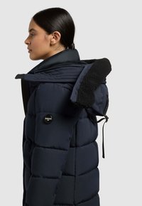 Marineblå polstret jakke med stor krave og sort fleecefór, mærke-knap og sidelomme med lynlås. Justerbar snøre i hætten.