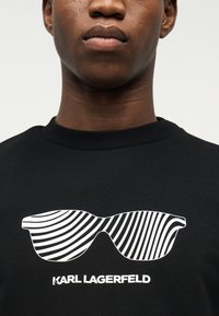 Άτομο που φοράει μαύρο πουκάμισο με γραφικό γυαλιών ηλίου με λευκές ρίγες και κείμενο «KARL LAGERFELD» κάτω από το σχέδιο.