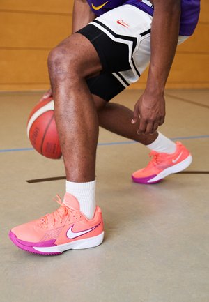 Baskets de basketball Nike aux couleurs vives rose et violet avec des détails en maille, arborant un logo blanc et des semelles texturées, sur un parquet.