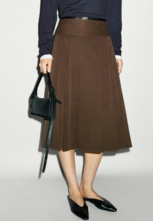 Personne portant une jupe midi plissée marron, des ballerines noires pointues et tenant un petit sac à main noir avec une longue bandoulière.