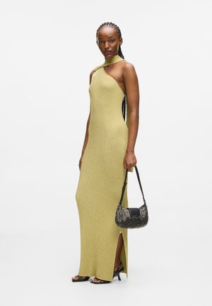Szydełkowa, bezrękawnikowa, długa żółta sukienka z wysokim dekoltem, z bocznymi rozcięciami, zestawiona z małą, czarną torebką z wypukłym detalem frędzli.