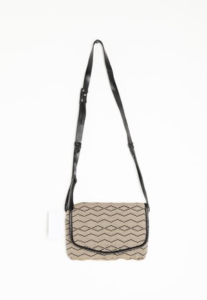 Sac bandoulière rectangulaire avec motif géométrique noir sur tissu beige et bandoulière en cuir noir réglable suspendu sur fond blanc.