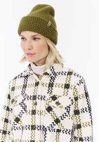 Mujer que lleva un gorro de punto verde oliva y una chaqueta crema con un patrón de cuadros en zigzag en verde oliva y negro sobre un suéter de cuello alto de canalé.