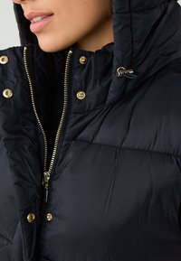 Giacca bomber blu navy con alto colletto, dettagli in metallo dorato, zip visibile e bottoni a pressione, con texture trapuntata e cappuccio.