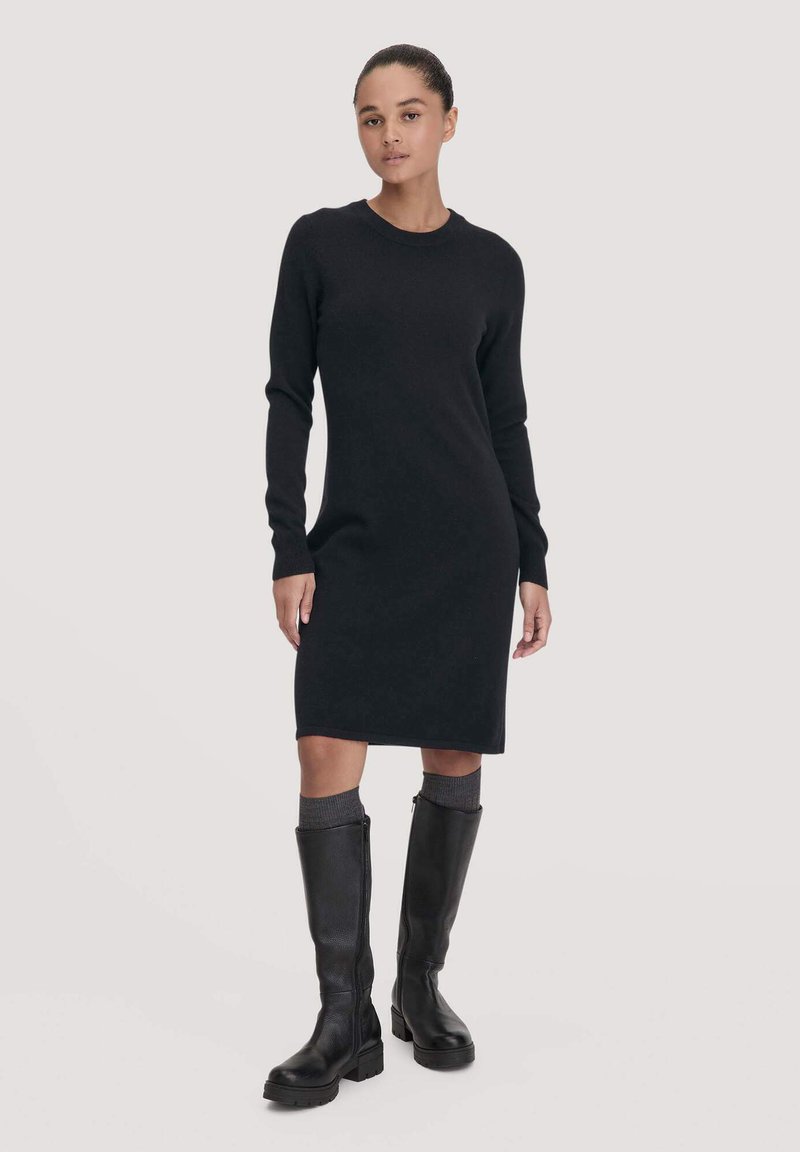 Schwarzes Strickkleid mit langen Ärmeln und Rundhalskragen, enganliegende Silhouette, kombiniert mit knielangen schwarzen Lederstiefeln und grauen Socken.