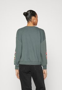Sweatshirt vert à col rond et épaules tombantes, orné de broderies florales colorées sur les manches, fabriqué en tissu doux.