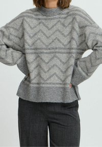 Personne portant un pull gris avec des motifs en zigzag et un ourlet côtelé, associé à un pantalon gris foncé, les mains posées sur les hanches.