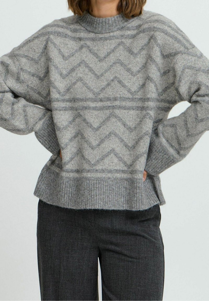 Personne portant un pull gris avec des motifs en zigzag et un ourlet côtelé, associé à un pantalon gris foncé, les mains posées sur les hanches.