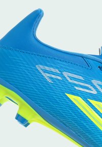 Gros plan sur une chaussure de football bleue avec une semelle et des crampons jaune néon, présentant une tige texturée et un marquage partiel blanc et jaune.