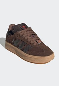 Braune Wildledersneaker mit schwarzen Akzenten, strukturierten grauen Streifen und einer Sohle aus Gummi. Schnürsenkel und Logo in kontrastierendem roten Detail.
