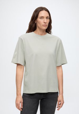 Kvinde med langt brunt hår iført en løs lysegrå T-shirt og sorte jeans, der står foran en ensfarvet hvid baggrund.
