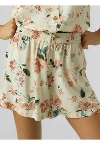 Nainen, jolla on päällänsä beige kukkakuosiset shortsit, joissa on elastinen nyörivyö, yhdistettynä samanlaiseen napitettuun kukkakuosiset paitaan.