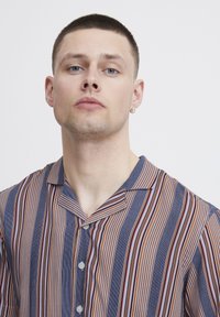 Camicia a maniche corte con righe verticali blu, rosse e bianche, realizzata in tessuto leggero. Presenta un colletto e una chiusura con bottoni.