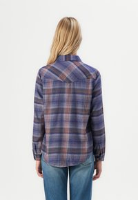 Woman with long blonde hair wearing a blue and purple plaid long-sleeve shirt and blue jeans, standing facing away against a white background.