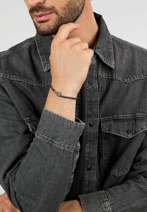 Camicia in denim nera con bottoni a pressione, indossata con un bracciale intrecciato scuro decorato con un accento metallico rotondo.