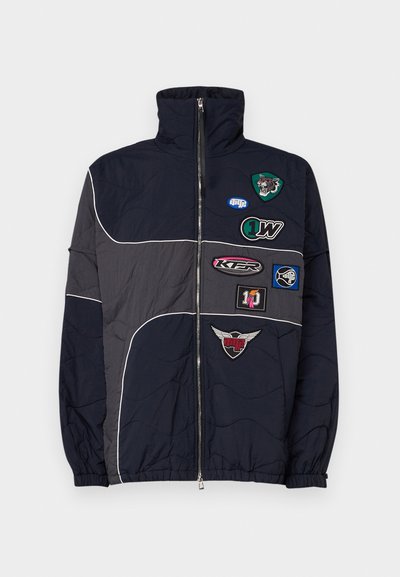 Navy and gray puffer jacket, zippered with high collar. Features multiple fabric patches with logos and embroidered accents on the chest.