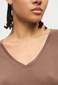 Κοντινό πλάνο του κάτω μέρους του προσώπου, του λαιμού και του πάνω μέρους του στήθους ενός ατόμου που φοράει καφέ μπλούζα με V-λαιμόκοψη και έχει κοντά πλεξούδες μαλλιών.