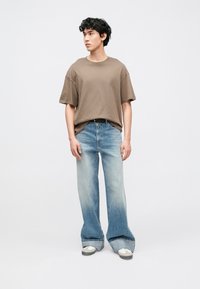 Braunes oversized T-Shirt kombiniert mit hellblauen, weit geschnittenen Jeans. Die Jeans weisen eine leichte Verwaschung und markante Saumdetails auf. Lässige Sneaker runden den Look ab.