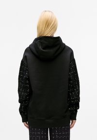 Black hoodie with textured black and white patterned sleeves, featuring a large hood and a relaxed fit. Smooth fabric on the body.