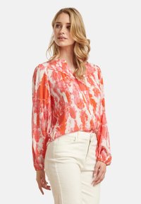 Seidenbluse mit einem roten und weißen abstrakten Muster, langen Ärmeln, Knopfverschluss und einem plissierten Kragen. Kombiniert mit weißen Jeans.