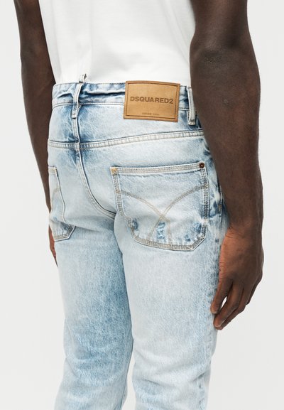 Homme portant un jean bleu clair déchiré avec un patch en cuir marron DSQUARED2 sur la ceinture arrière, une chemise blanche et un bras à peau foncée visible.