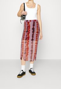 Femme portant un débardeur blanc et une jupe midi tie-dye semi-transparente, avec des baskets noires et des chaussettes blanches, tenant un sac à bandoulière noir.