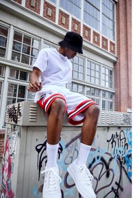Camiseta blanca, pantalones cortos de baloncesto blancos con acentos rojos, sombrero de cubo negro, zapatillas blancas, sentado sobre una caja de servicios cubierta de graffiti.