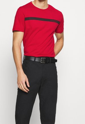 Man wearing a red short-sleeve t-shirt with a thin black stripe across the chest, black belt, and black pants, standing against white background.