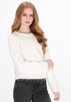 Ung kvinde med langt bølget blondt hår iført en hvid strikket sweater og sorte jeans, poserende mod en ensfarvet hvid baggrund.