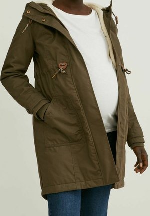 Zwangere persoon die een open olijfgroene parka met beige voering draagt, een wit shirt en een blauwe spijkerbroek, met één hand in de zak.