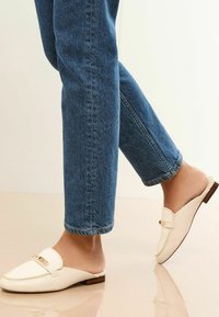 Witte leren instaploafers met een gouden accent, gecombineerd met blauwe denim jeans. De schoen heeft een lage hak en een gladde textuur.