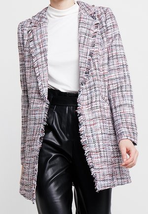 Personne portant un col roulé blanc, un pantalon en cuir noir taille haute et une longue veste en tweed multicolore avec des bords effrangés.