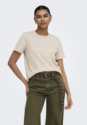 Mujer con cabello oscuro, camisa beige con texto punteado y pantalones verde oliva, que lleva un cinturón marrón de eslabones, de pie con una mano en la cadera.