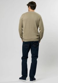 Beige, hosszú ujjú pulóver ribbelt mintával, sötétkék farmernadrággal, amelynek különleges zsebkialakítása van, és fehér sportcipővel.