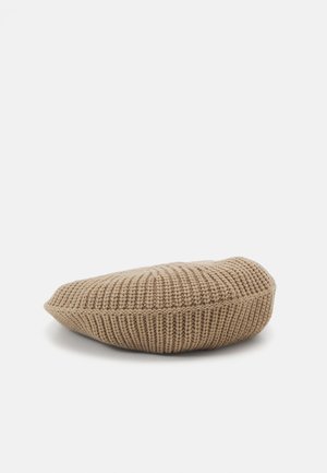Béret beige en tricot avec texture côtelée, couronne plate et forme ronde et douce reposant sur une surface blanche.