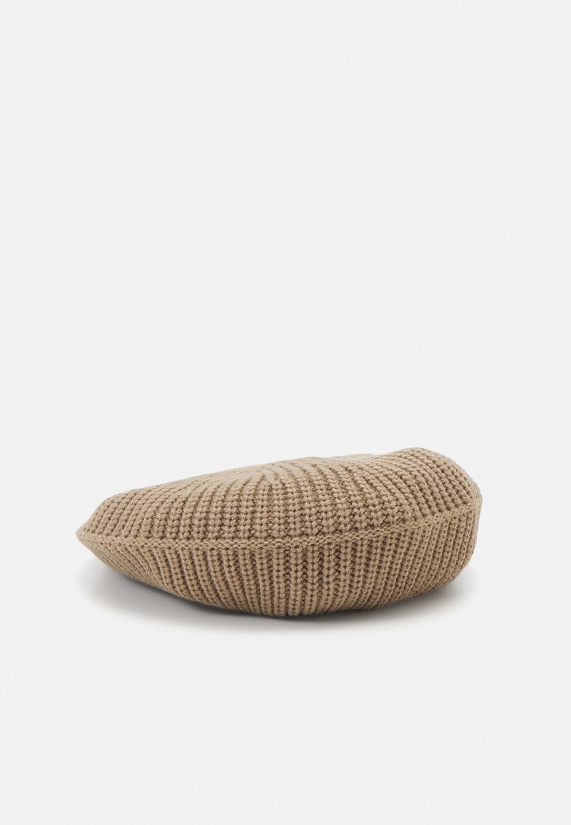 Béret beige en tricot avec texture côtelée, couronne plate et forme ronde et douce reposant sur une surface blanche.