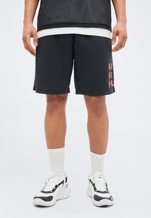 Mężczyzna w czarnych sportowych szortach z pomarańczowymi logo Under Armour, białych skarpetach sięgających do kostek oraz białych i czarnych sneakersach, stojący na jednolitym tle.