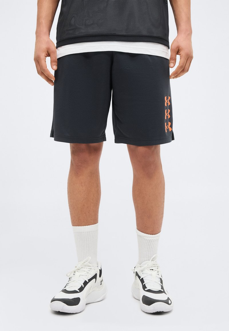 Άνδρας που φοράει μαύρα αθλητικά σορτς με πορτοκαλί λογότυπα Under Armour, λευκές κάλτσες crew και λευκά και μαύρα αθλητικά παπούτσια, στέκεται μπροστά σε ένα απλό φόντο.