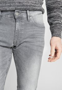 Sivé džínsy s úzkym strihom, päť vreciek, zapínanie na gombík a jemne vyblednutá textúra. Nosené s pleteným svetríkom s vzorom.