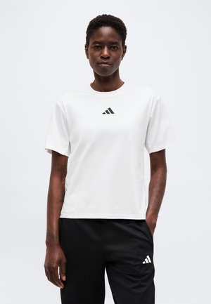 Osoba nosící bílé tričko Adidas a černé kalhoty Adidas stojí s jednou rukou v kapse před jednobarevným pozadím.